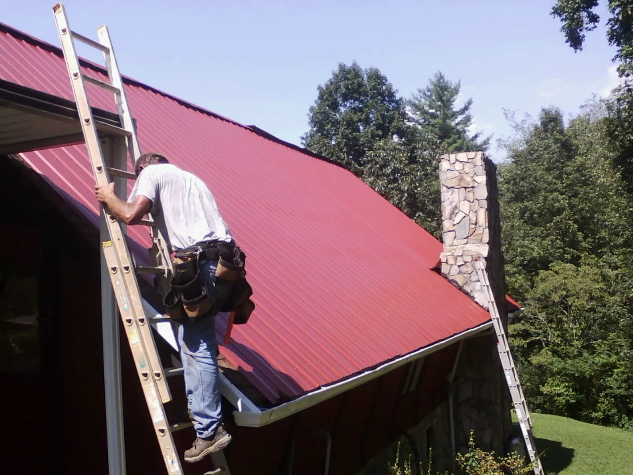 Roofer on a ladder beside a red metal roof and stone chimney — Soffit & Fascia Repair by Roofing Brothers in Walterboro